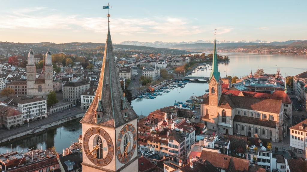 Aerial photograph of Zurich, showing Lake Zurich and the nearby alps in the background, with the Limmat, parts of the old town and prominently the three chruchers, Grossmuenster, Fraumuenster and St. Peter in the foreground.